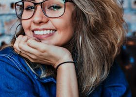 A young woman wearing glasses and denim, smiling in an indoor setting.